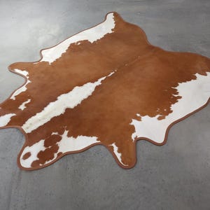 May include: A cowhide rug with a natural brown and white pattern. The rug has a unique shape with a brown border. The rug is a decorative floor covering, adding a rustic touch to any room.