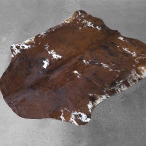 May include: A natural cowhide rug featuring a large, irregular shape with a primary deep brown color and scattered patches of white fur. The hide displays a smooth, flat texture with a distinct organic outline and varied dark brown markings.