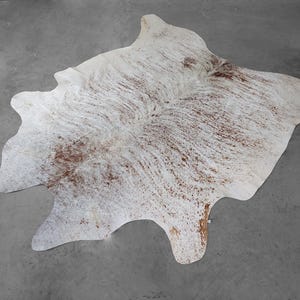 May include: A natural cowhide rug featuring an irregular, organic shape with a white base color and scattered brown speckled patterns throughout the surface. The hide displays a textured hair-on-hide finish with distinct brown markings.