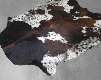May include: A natural cowhide rug featuring an irregular, organic shape with a tri-color pattern of dark brown, black, and white patches. The hide displays a mix of solid dark sections and speckled areas, showcasing the unique texture of the animal fur.