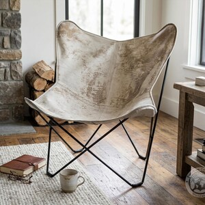May include: A butterfly chair featuring a white and tan cowhide seat with a distressed, mottled pattern. The seat is supported by a thin, black metal frame with a cross-base design. The chair has a wide, curved backrest and a deep, sloped seat.