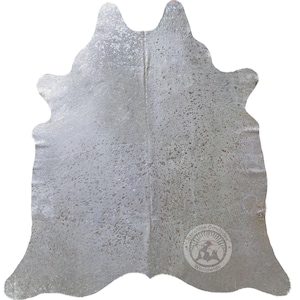 May include: A silver-toned cowhide rug featuring a metallic finish with a natural speckled pattern throughout the surface. The hide has an irregular, organic shape and includes a circular logo stamp in the bottom right corner reading Sunshine Cowhides Worldwide.