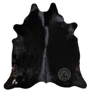 May include: A solid black cowhide rug with a natural, irregular organic shape. The surface features a smooth, glossy texture throughout. A circular logo with the text Sunshine Cowhides Worldwide is visible in the bottom right corner of the hide.
