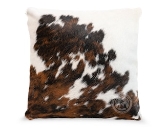 May include: A square throw pillow featuring a natural cowhide pattern with patches of white, dark brown, and black fur. The front surface displays a circular logo stamp in the bottom right corner with the text Cowhide Worldwide.