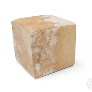 May include: A square ottoman upholstered in genuine cowhide featuring a natural pattern of light tan and white patches. The cube-shaped furniture piece has a textured hair-on-hide surface with visible variations in colour across all visible sides.