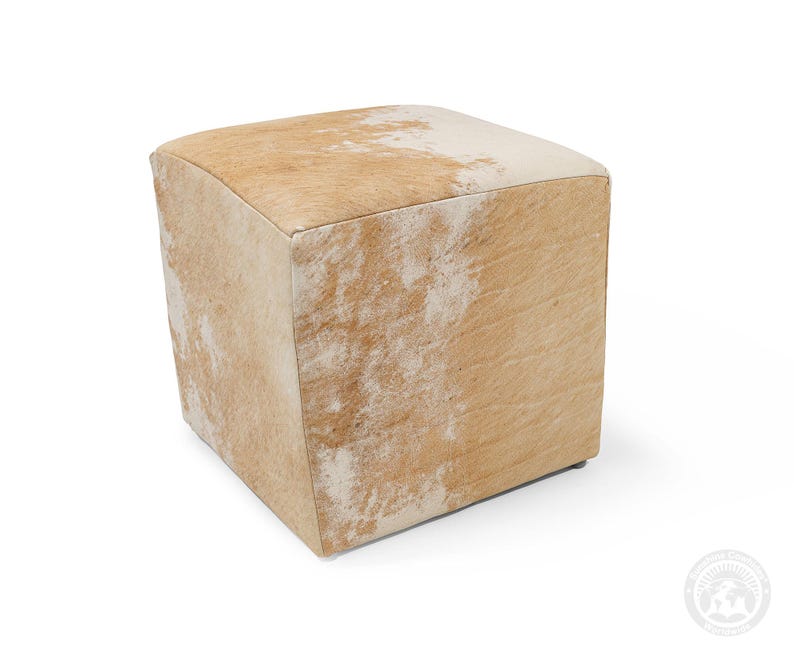 May include: A square ottoman upholstered in genuine cowhide featuring a natural pattern of light tan and white patches. The cube-shaped furniture piece has a textured hair-on-hide surface with visible variations in colour across all visible sides.