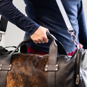 May include: A black duffel bag featuring a central panel with a brown and black mottled animal print pattern. The bag includes black leather-like handles, silver-toned metal rivets, a detachable shoulder strap with metal clips, and side zippers.