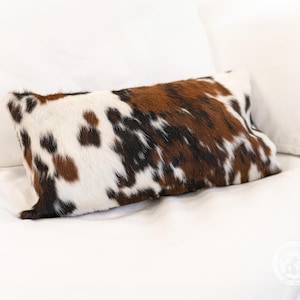 May include: A rectangular throw pillow featuring a natural cowhide pattern with a mix of white, brown, and black patches. The surface has a textured, hair-on-hide appearance with irregular spots distributed across the entire front of the pillow.