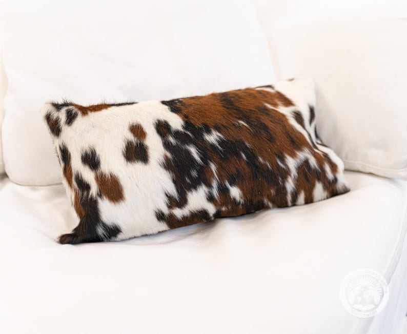 May include: A rectangular throw pillow featuring a natural cowhide pattern with a mix of white, brown, and black patches. The surface has a textured, hair-on-hide appearance with irregular spots distributed across the entire front of the pillow.