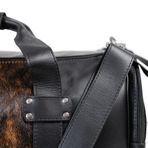 May include: A black leather duffel bag featuring a brown and black animal print hair-on hide panel. The bag includes a sturdy top handle, a detachable shoulder strap, silver-toned metal rivets, and a black zipper closure along the side.