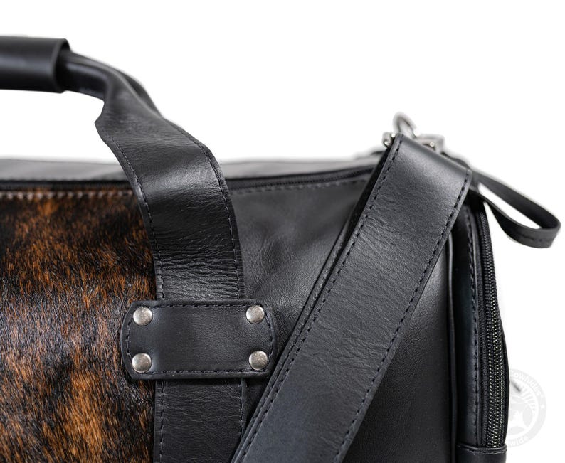 May include: A black leather duffel bag featuring a brown and black animal print hair-on hide panel. The bag includes a sturdy top handle, a detachable shoulder strap, silver-toned metal rivets, and a black zipper closure along the side.