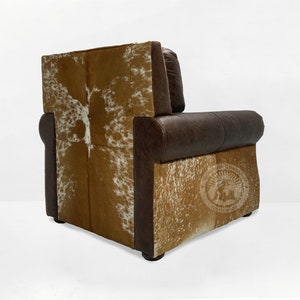 May include: A brown leather armchair with a cowhide back and seat. The chair has a tufted back and a rolled armrest. The chair is sitting on a white background.