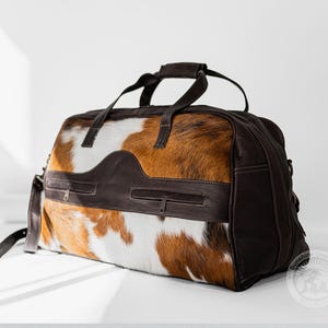 May include: A duffel bag featuring a brown and white cowhide pattern with dark brown leather accents. The bag includes two zippered pockets on the front, two top handles, and a side strap. A circular logo with the text Sunshine Cowhide is visible.