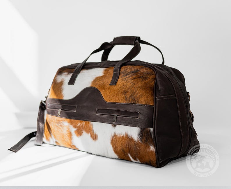 May include: A duffel bag featuring a brown and white cowhide pattern with dark brown leather accents. The bag includes two zippered pockets on the front, two top handles, and a side strap. A circular logo with the text Sunshine Cowhide is visible.