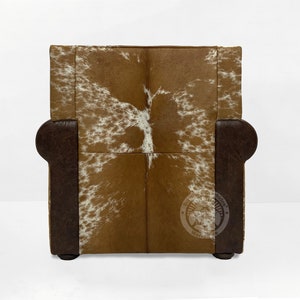 May include: A brown leather armchair with a white and brown cowhide pattern on the back and seat. The chair has a dark brown wooden frame and legs.