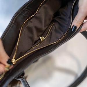 May include: A brown leather tote bag with a gold zipper. The bag is open and the inside is lined with a brown fabric.