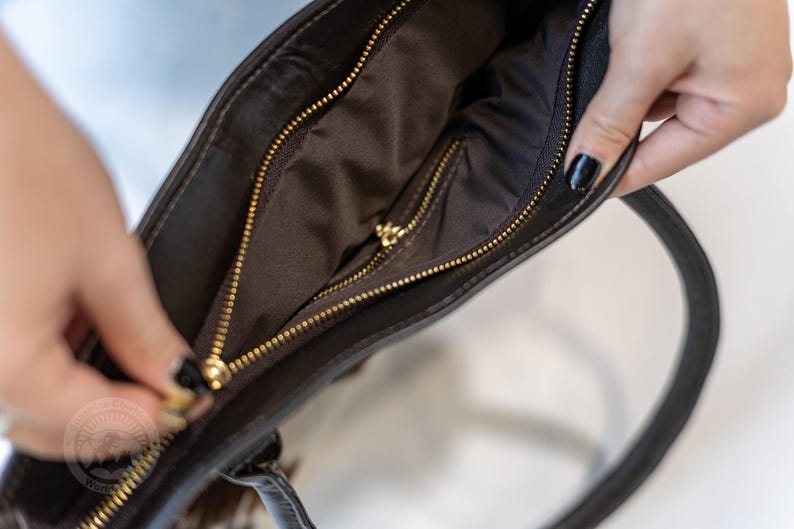 May include: A brown leather tote bag with a gold zipper. The bag is open and the inside is lined with a brown fabric.