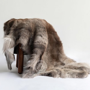 May include: A brown and grey fur rug draped over a wooden stool. The rug has a soft, textured appearance with varying shades of brown and grey. The stool has three dark wooden legs. The rug is on a white background.