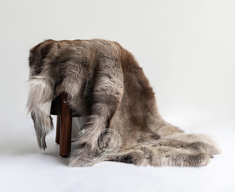May include: A brown and grey fur rug draped over a wooden stool. The rug has a soft, textured appearance with varying shades of brown and grey. The stool has three dark wooden legs. The rug is on a white background.