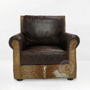 May include: A brown leather armchair with a cowhide base and gold nailhead trim. The chair has a tufted back and rolled arms. The chair is made by the "Machine Co."
