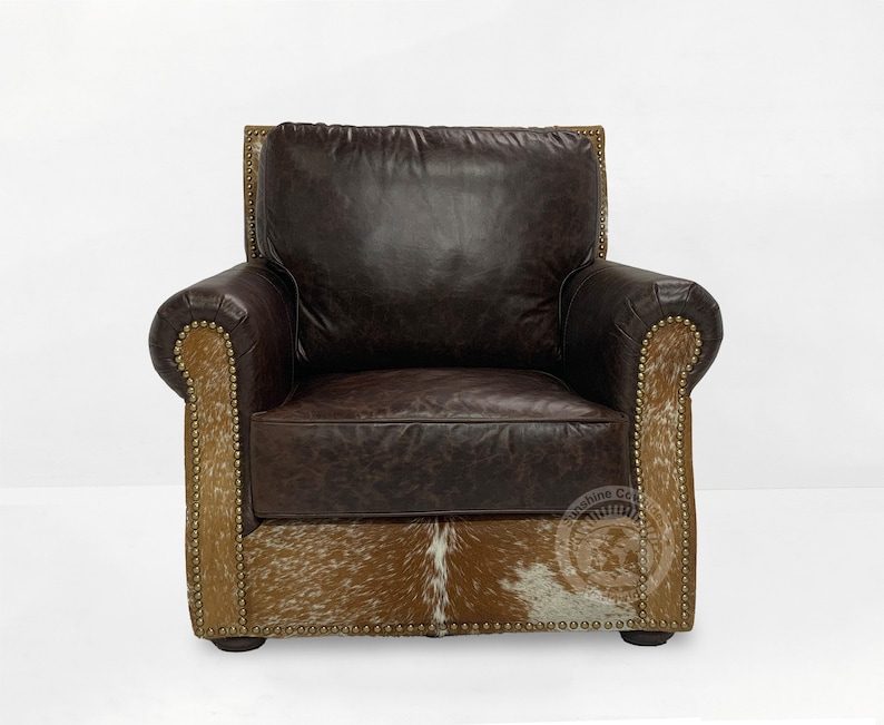 May include: A brown leather armchair with a cowhide base and gold nailhead trim. The chair has a tufted back and rolled arms. The chair is made by the "Machine Co."