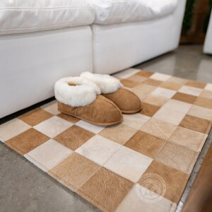 May include: A pair of brown suede slippers with white faux fur lining, placed on a brown and white checkered cowhide rug.