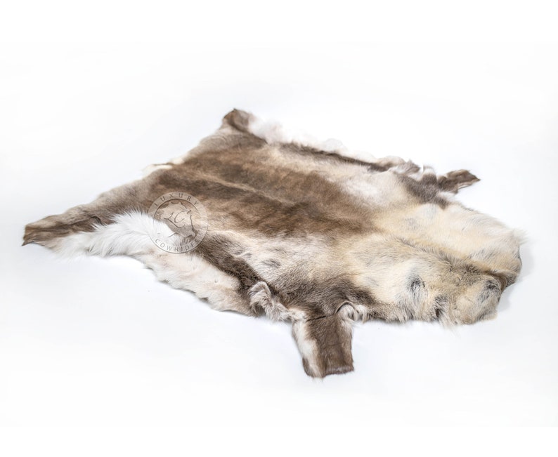 May include: A reindeer hide rug in natural shades of brown, grey, and white. The fur is thick and soft, with variations in tone and texture. The hide is irregularly shaped, with a natural edge.