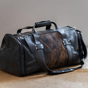 May include: A black duffel bag featuring a central panel of brown and black cowhide hair-on leather. The bag includes black leather handles, a detachable shoulder strap with a padded section, silver-toned metal hardware, and a side zipper pocket.