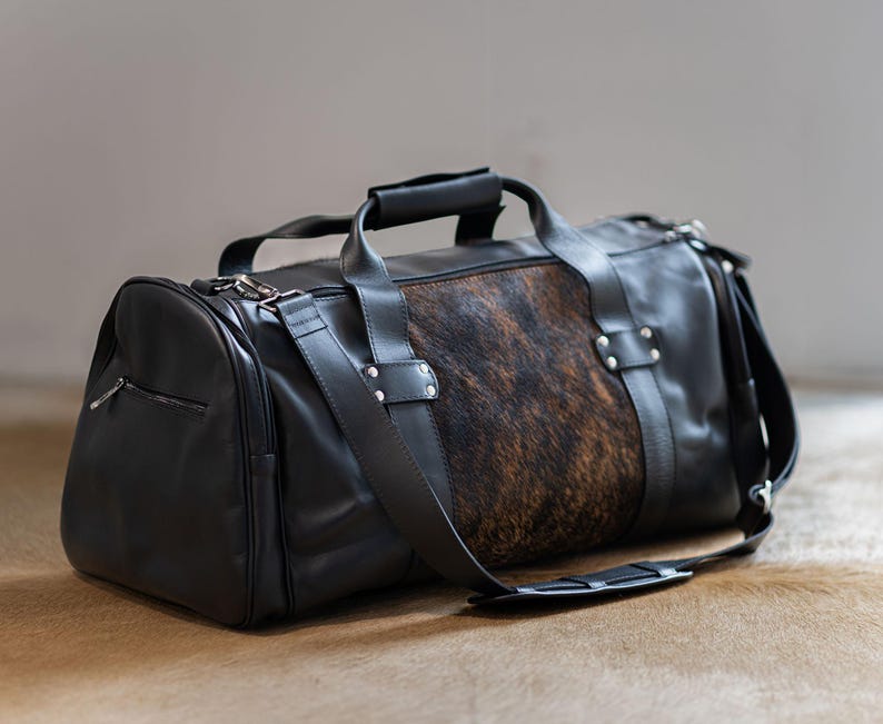 May include: A black duffel bag featuring a central panel of brown and black cowhide hair-on leather. The bag includes black leather handles, a detachable shoulder strap with a padded section, silver-toned metal hardware, and a side zipper pocket.