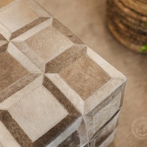 Pu&ograve; includere: Un pouf quadrato con un motivo geometrico in pelle di mucca marrone chiaro e bianco. Il pouf &egrave; realizzato con un patchwork di diversi pezzi di pelle, creando un design unico ed elegante.