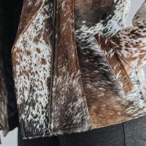 May include: A cowhide jacket with a zipper closure. The jacket features a mix of brown, white, and black patches. The jacket has a pocket on the front and a zipper closure. The jacket is made of leather.