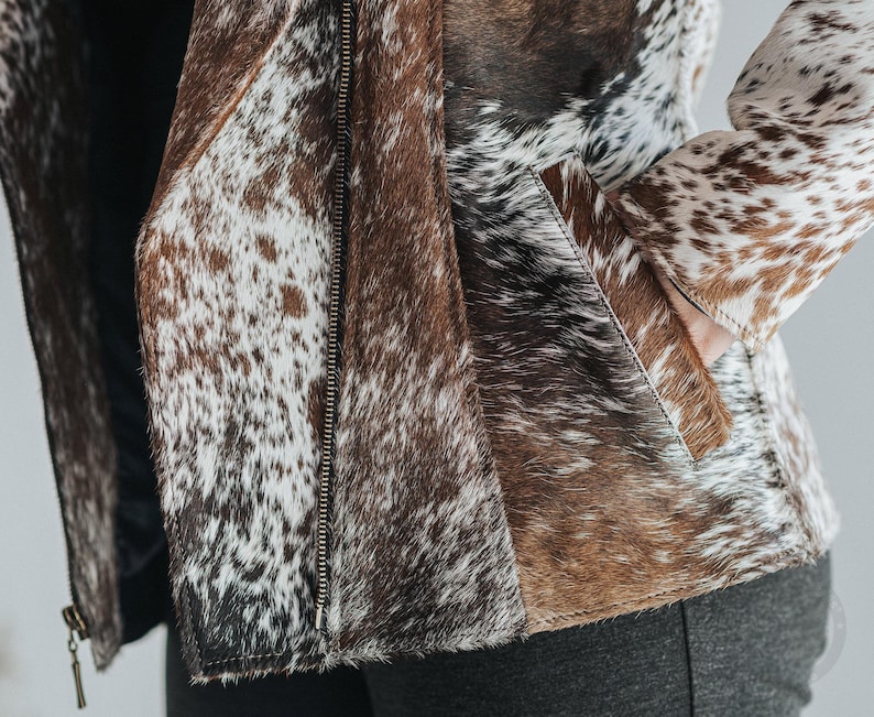 May include: A cowhide jacket with a zipper closure. The jacket features a mix of brown, white, and black patches. The jacket has a pocket on the front and a zipper closure. The jacket is made of leather.