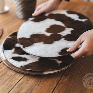 Op de afbeelding: Twee bruine en witte koeienhuid placemats op elkaar gestapeld. De placemats zijn rond en hebben een natuurlijke, rustieke uitstraling.
