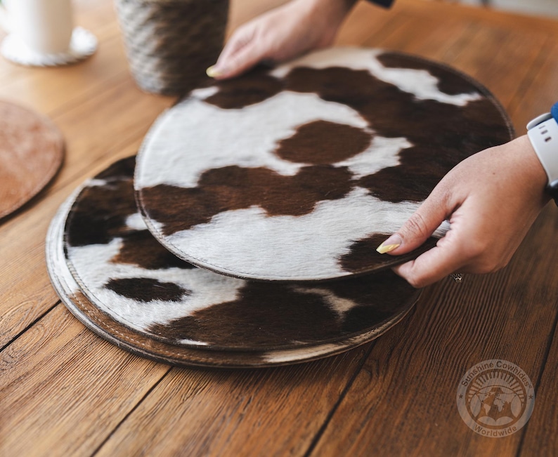 May include: Two round placemats featuring a brown and white cowhide pattern with a smooth texture. The circular mats have a dark brown stitched edge and a natural animal print design. A circular logo with the text Sunshine Cowhides Worldwide is visible.