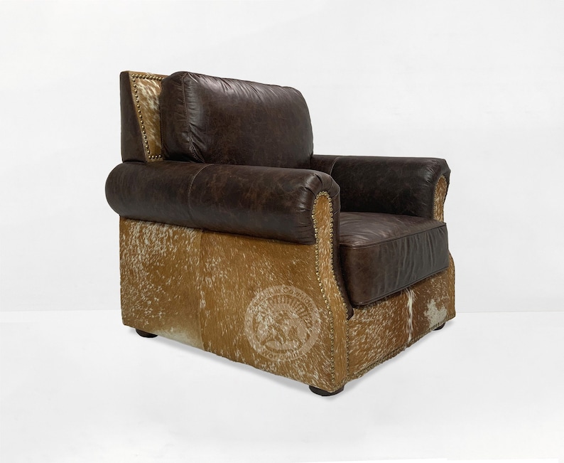 May include: A brown leather armchair with a cowhide base. The chair has a tufted back and a rolled armrest. The chair is accented with brass nailhead trim.