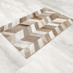 Pode incluir: Um tapete de couro com padr&atilde;o chevron marrom e branco. O tapete est&aacute; colocado em um piso de madeira clara.