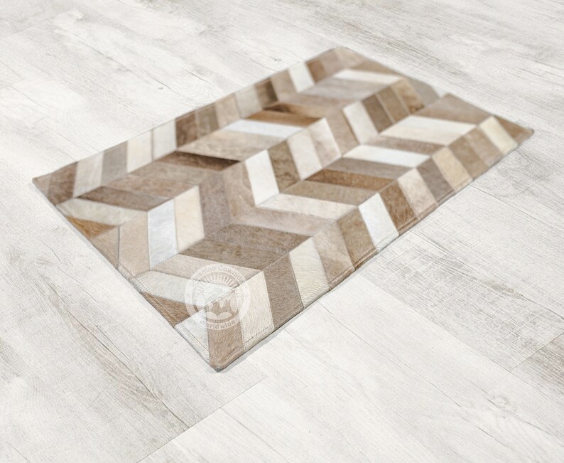 Pode incluir: Um tapete de couro com padr&atilde;o chevron marrom e branco. O tapete est&aacute; colocado em um piso de madeira clara.