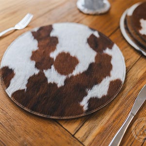May include: A round placemat featuring a natural cowhide pattern with patches of brown and white fur. The circular mat has a stitched edge and a flat profile, displaying a textured hair-on-hide surface with irregular spots across the entire top.