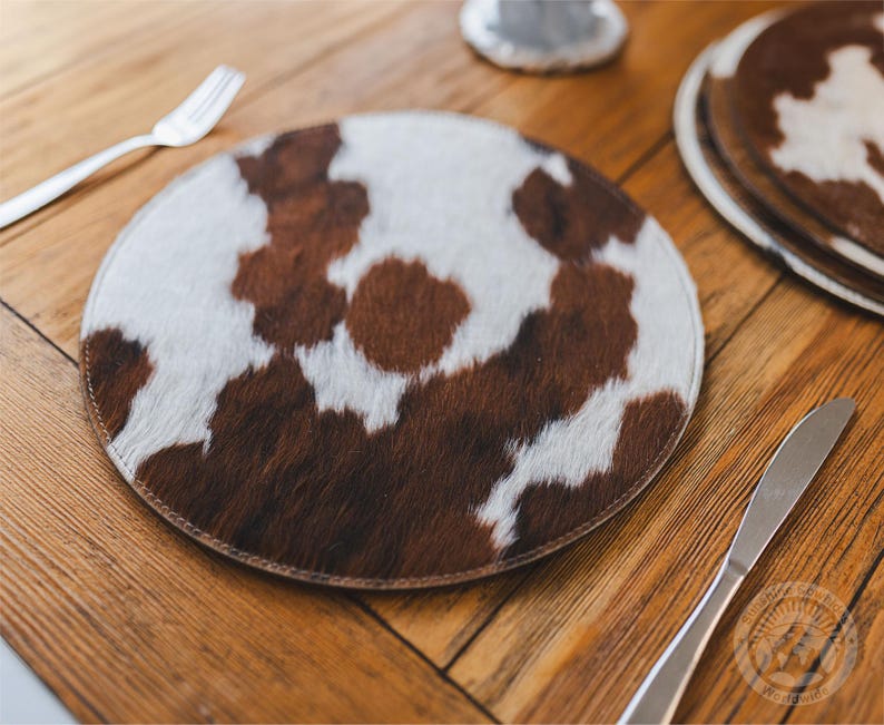 May include: A round placemat featuring a natural cowhide pattern with patches of brown and white fur. The circular mat has a stitched edge and a flat profile, displaying a textured hair-on-hide surface with irregular spots across the entire top.