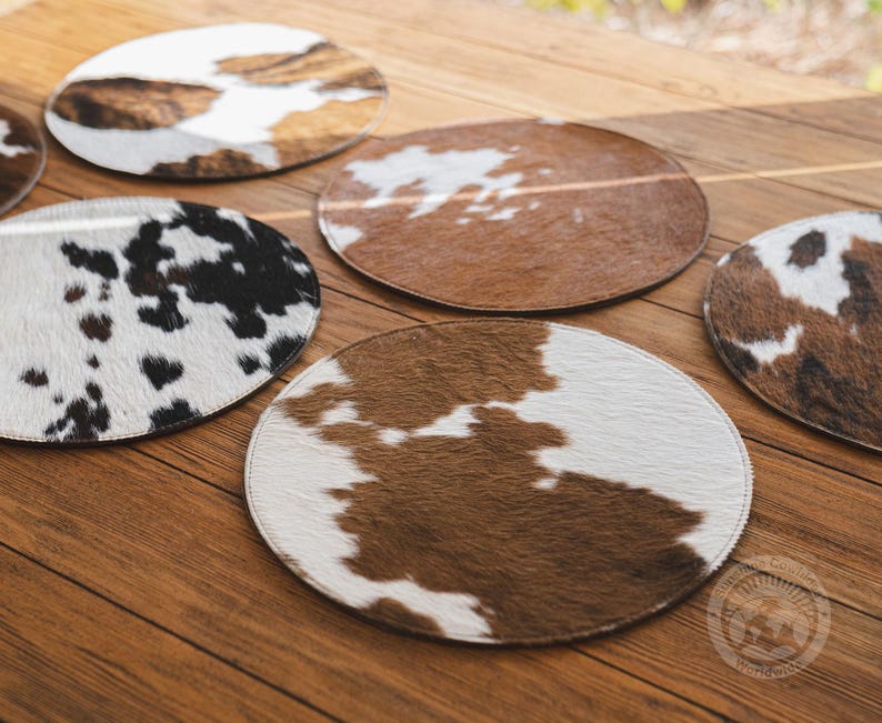 May include: A collection of circular coasters featuring various cowhide patterns in shades of brown, white, and black. Each coaster has a distinct animal print texture with stitched edges, displaying natural color variations across the set.