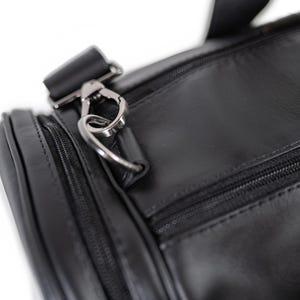 May include: A black duffel bag featuring a smooth surface, a zippered main compartment, and a side pocket. The bag includes a silver-toned metal lobster clasp attached to a D-ring, which connects to a black strap with matching metal hardware.