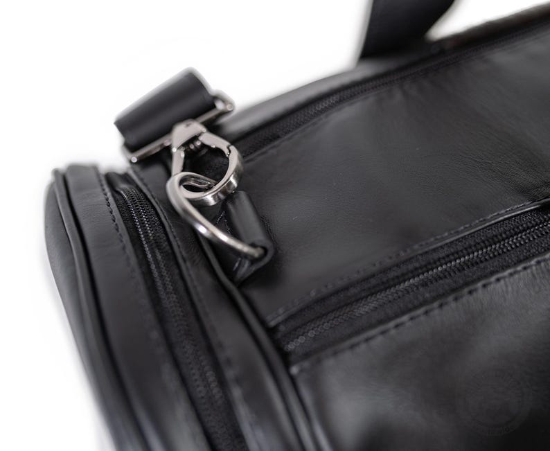 May include: A black duffel bag featuring a smooth surface, a zippered main compartment, and a side pocket. The bag includes a silver-toned metal lobster clasp attached to a D-ring, which connects to a black strap with matching metal hardware.