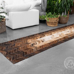 May include: A long, rectangular cowhide rug with a geometric pattern in shades of brown and tan. The rug features a herringbone design on the edges and a lighter, more natural-toned center. The rug is placed on a gray concrete floor.