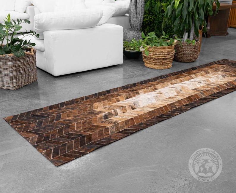 May include: A long, rectangular cowhide rug with a geometric pattern in shades of brown and tan. The rug features a herringbone design on the edges and a lighter, more natural-toned center. The rug is placed on a gray concrete floor.