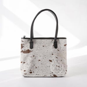 May include: A white and brown cowhide tote bag with black leather handles and trim.