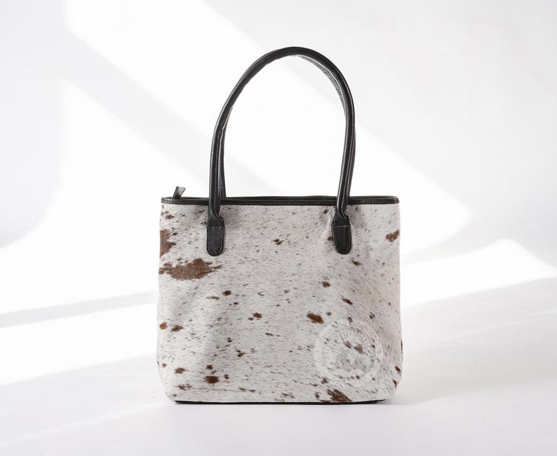 May include: A white and brown cowhide tote bag with black leather handles and trim.