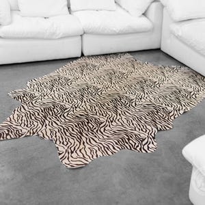 May include: A black and white zebra print cowhide rug with a cream background. The rug is laid out on a gray floor in front of a white sofa.