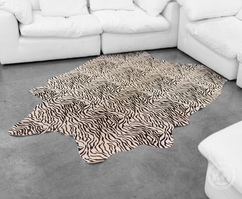 May include: A black and white zebra print cowhide rug with a cream background. The rug is laid out on a gray floor in front of a white sofa.