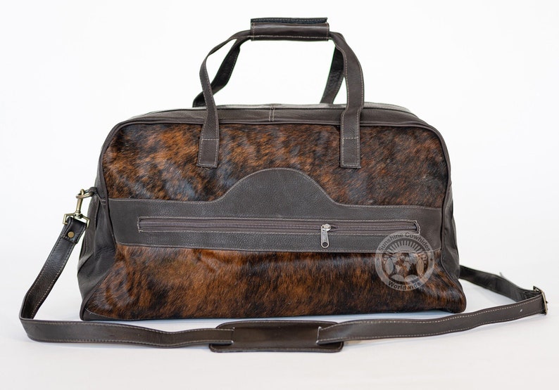 May include: A brown and black cowhide duffel bag featuring smooth dark brown leather trim, dual top handles, and an adjustable shoulder strap. The front includes a horizontal zippered pocket and a circular logo with the text Sunshine Cowhide Worldwide.