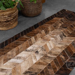 May include: A rectangular rug with a chevron pattern, featuring various shades of brown and tan. The rug is made of a natural material, possibly cowhide, and has a textured appearance. The design creates a visual flow across the surface.